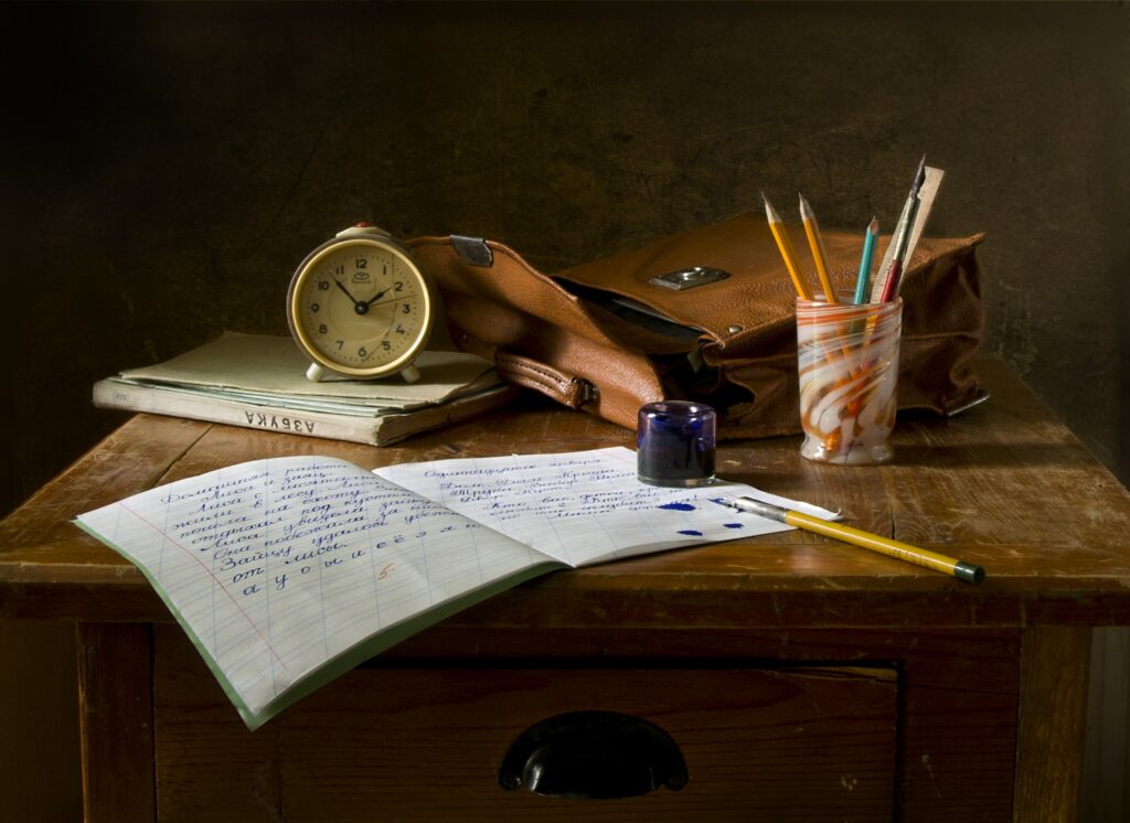 Photo of small wooden desk with messenger bag, desk clock, cup of pencils, and notebooks. One note book is open and has illegible writing.